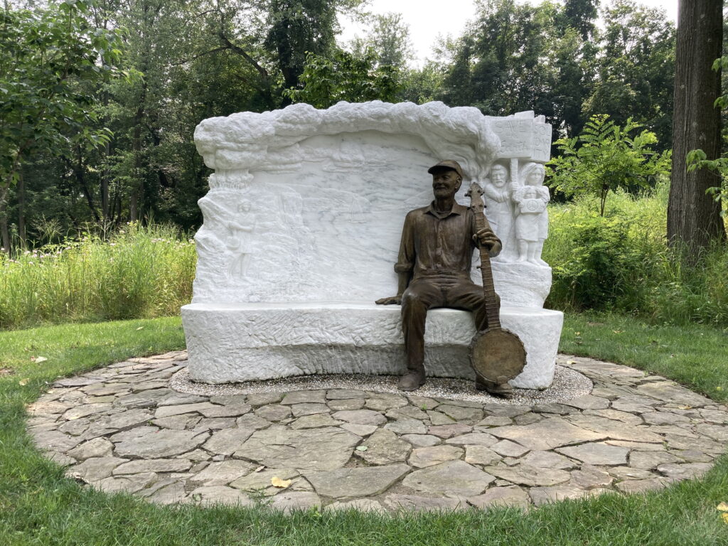 Over Life-size bronze portrait figure of Pete Seeger sitting with his banjo on 23 tons of Danby White Imperial Marble, carving including, protestors, slogans, environmental representation of the Clearwater Sloop on the Hudson River, located at the Puffin Foundation, in Teaneck NJ. Completed by artist G.L. Sussman in 2018.